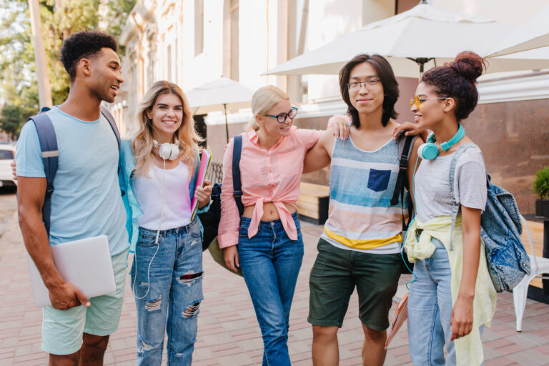 High school students walk between buildings on a college campus carrying backpacks and notebooks.
